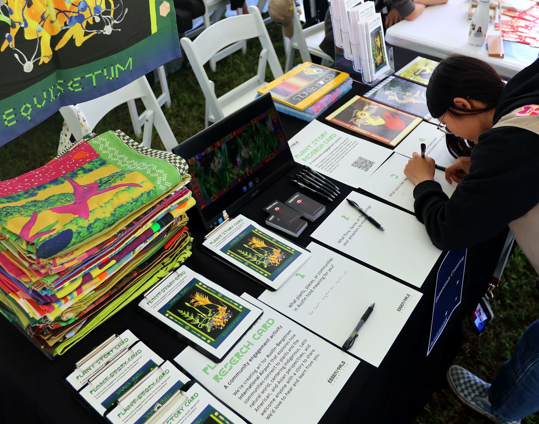 A participant writing their answers on a Plant Story Card at a table spread with printed cards and stacked colored paper.